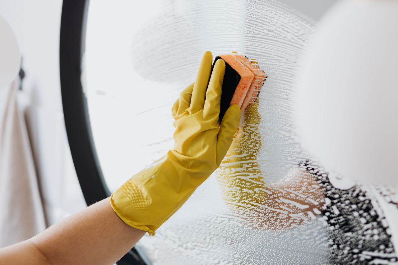 about-us-01 Anonymous person in yellow rubber gloves using simple sponge for cleaning mirror in bathroom